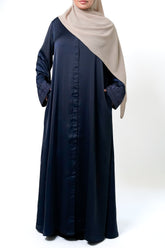 A woman wearing a navy blue button-down abaya with intricate embroidery on the sleeves. She is also wearing a beige hijab and has her hands in the pockets of the abaya