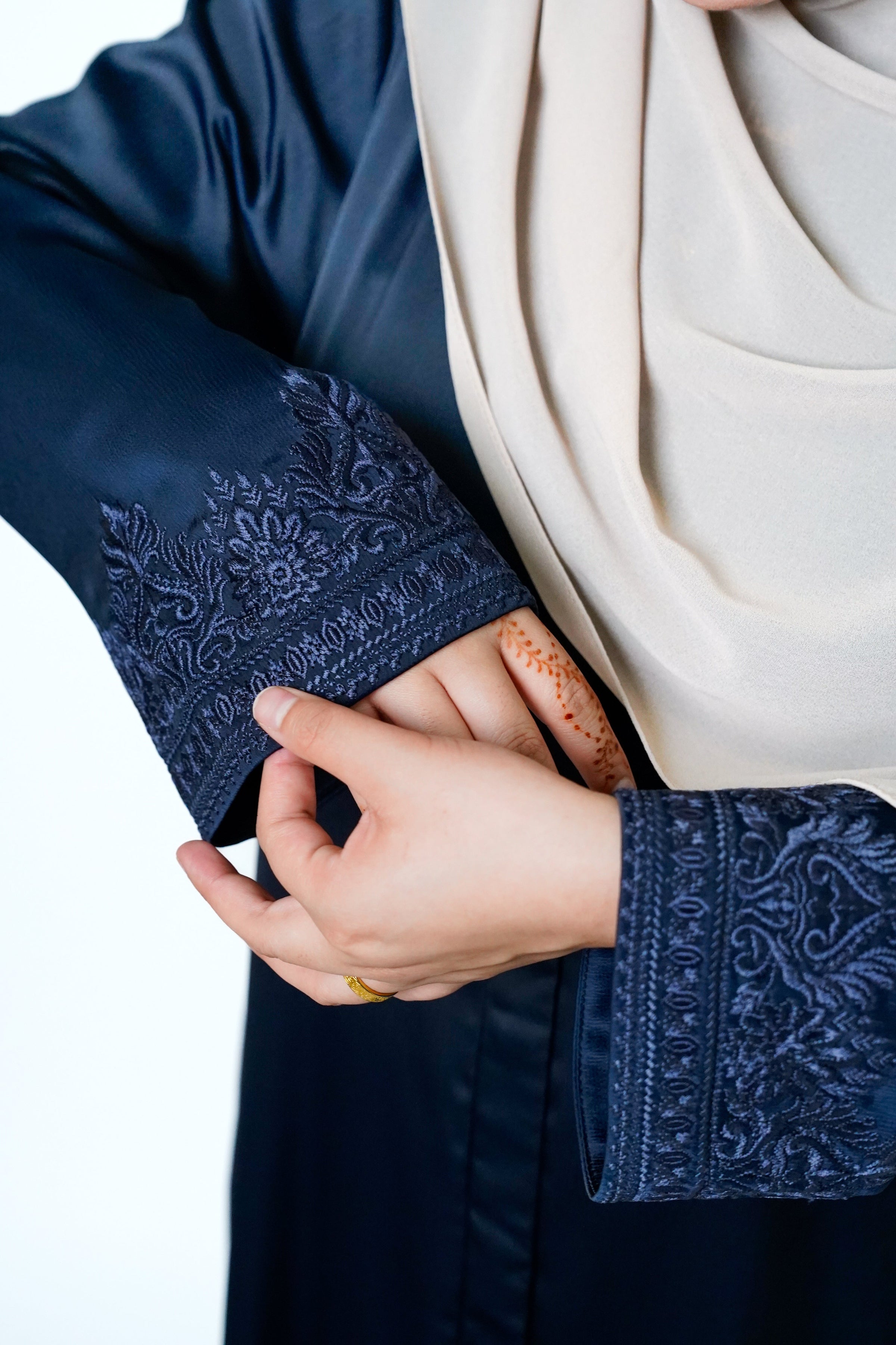 Close-up of a woman's hand adjusting the sleeve of a navy blue abaya with intricate dark embroidery on the cuffs. The woman is wearing a beige hijab.