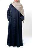 A woman wearing a navy blue button-down abaya with intricate embroidery on the sleeves. She is also wearing a beige hijab and has her hands in the pockets of the abaya