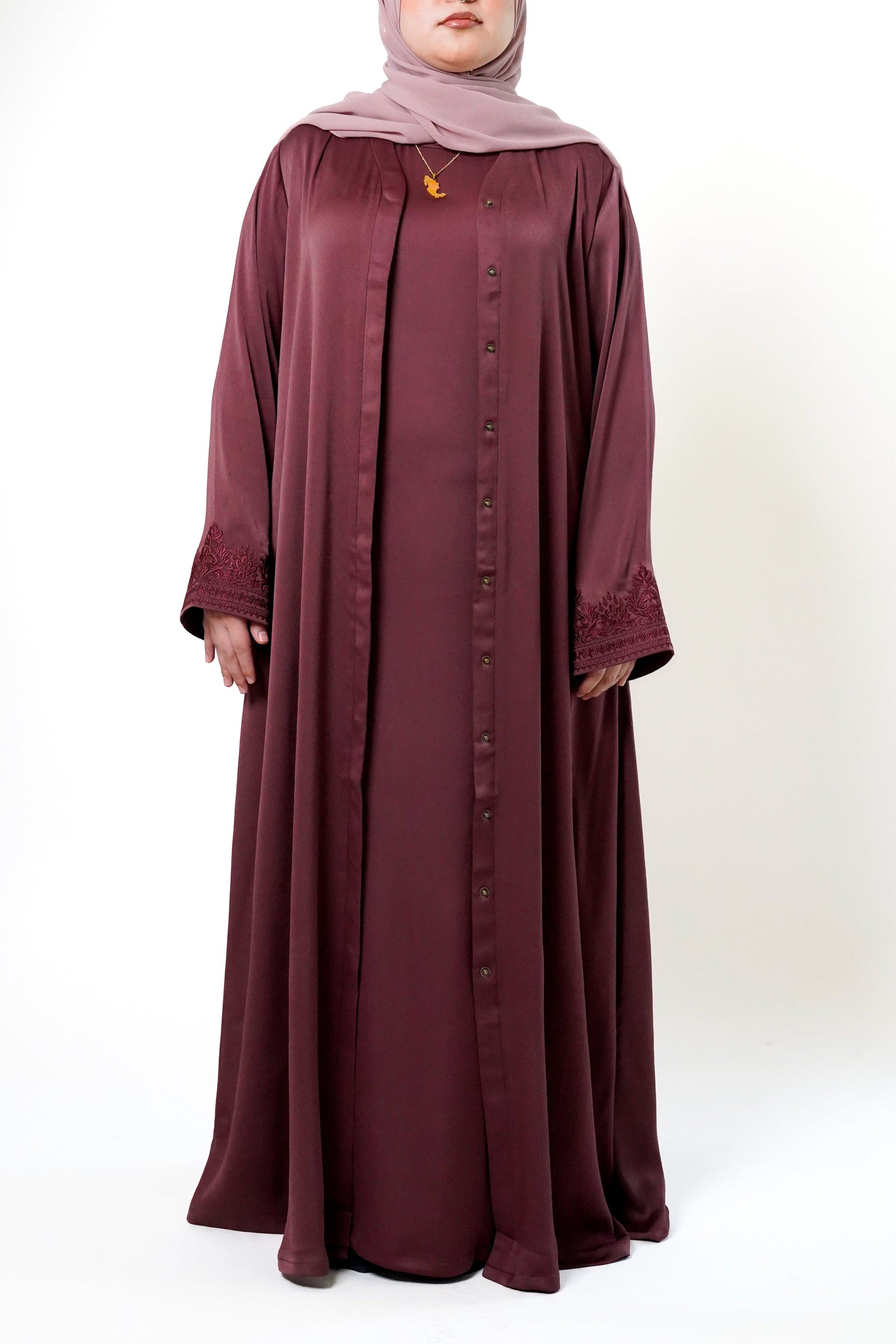 A woman wearing a maroon-colored abaya with a button-down front and intricate floral embroidery on the sleeves. She is also wearing a light pink hijab and a necklace, with her hands in the pockets of the abaya