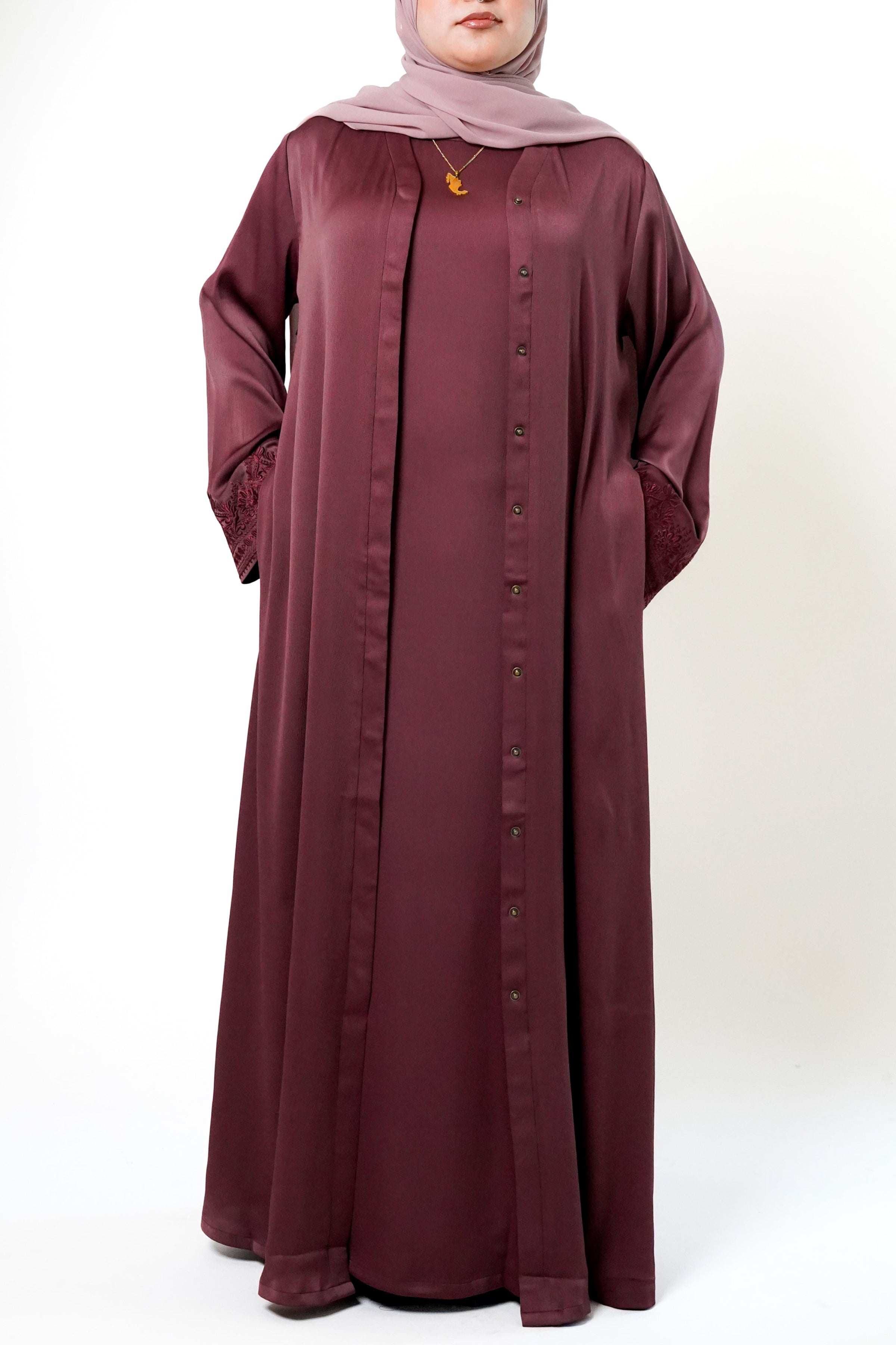 A woman wearing a maroon-colored abaya with a button-down front and intricate floral embroidery on the sleeves. She is also wearing a light pink hijab and a necklace, with her hands in the pockets of the abaya