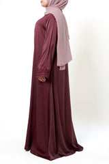 A woman wearing a maroon-colored abaya with a button-down front and intricate floral embroidery on the sleeves. She is also wearing a light pink hijab and a necklace, with her hands in the pockets of the abaya