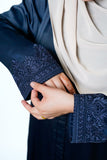 Close-up of a woman's hand adjusting the sleeve of a navy blue abaya with intricate dark embroidery on the cuffs. The woman is wearing a beige hijab.