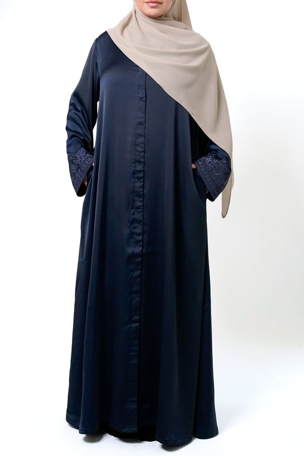 A woman wearing a navy blue button-down abaya with intricate embroidery on the sleeves. She is also wearing a beige hijab and has her hands in the pockets of the abaya